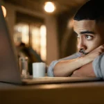 Homem pensativo olhando para a tela do laptop em um ambiente acolhedor e bem iluminado, com expressão de reflexão ou cansaço.