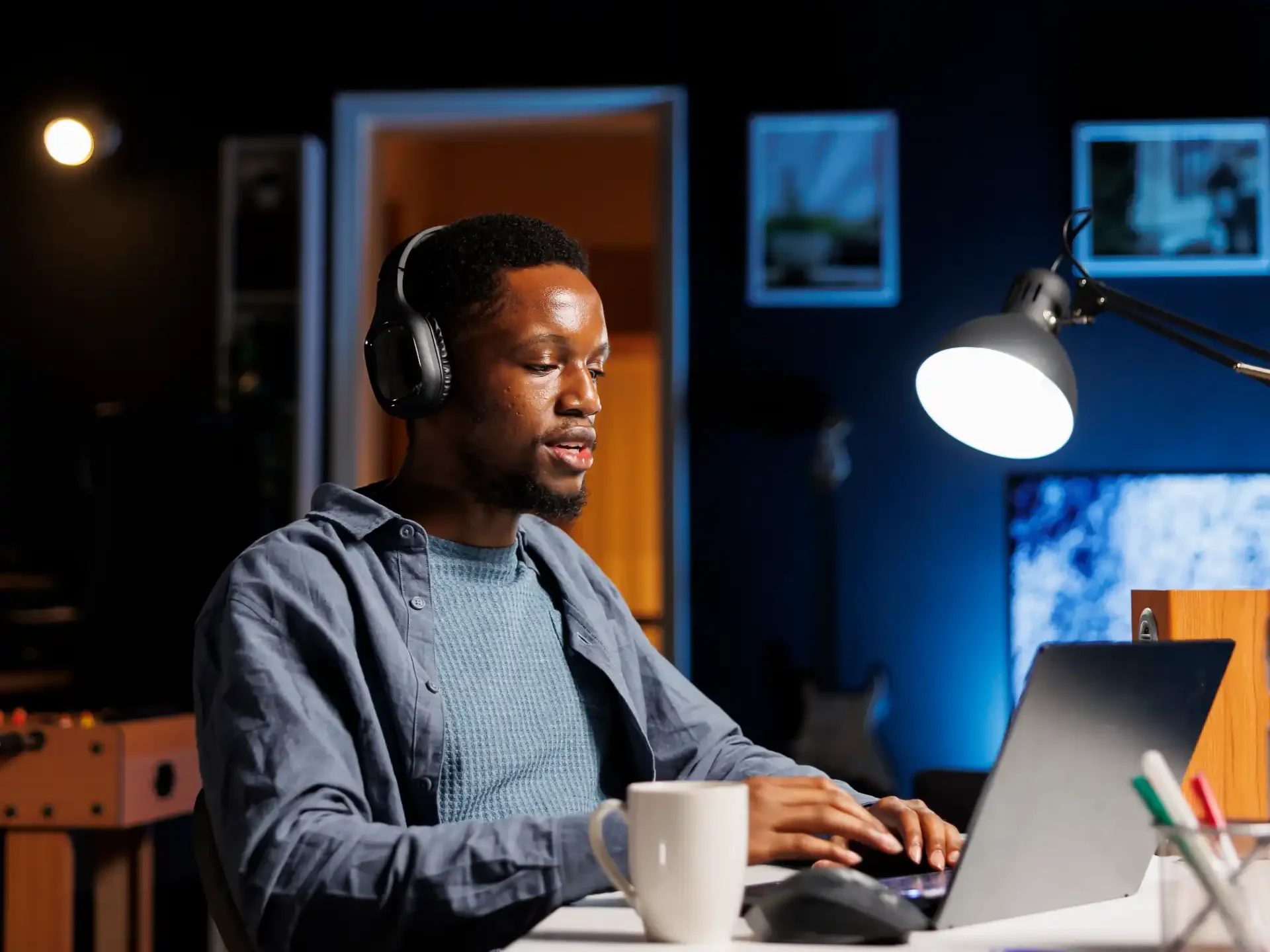 Homem usando fones de ouvido e trabalhando em um laptop em um ambiente moderno e aconchegante, com ilumina&ccedil;&atilde;o suave e decora&ccedil;&atilde;o elegante.