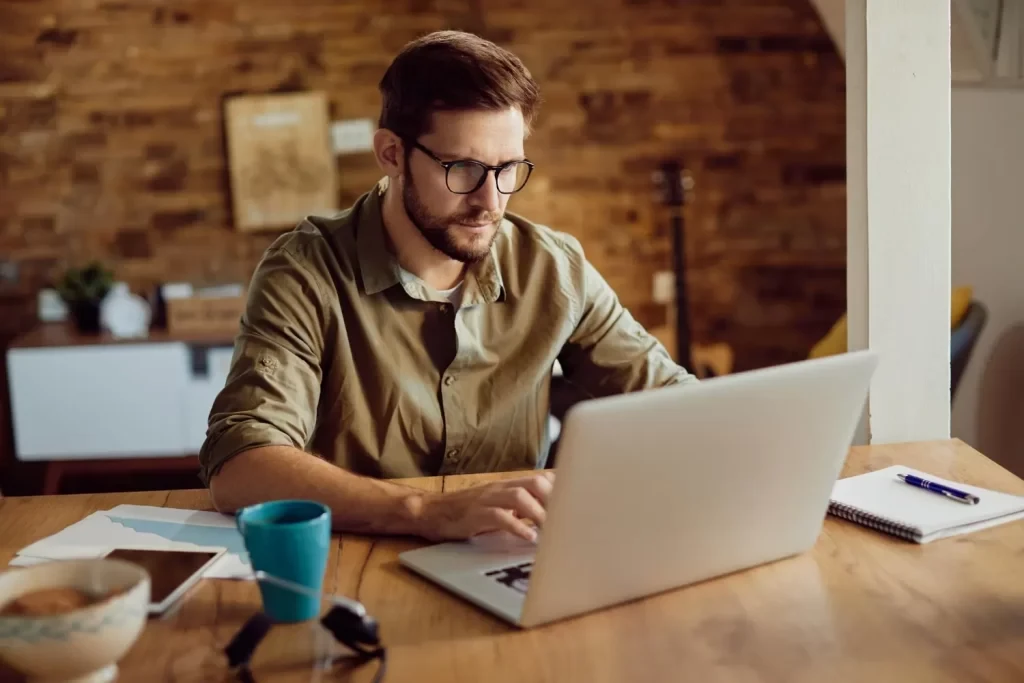 Homem de &oacute;culos escrevendo em seu laptop em uma mesa de madeira, ambiente de coworking com decora&ccedil;&atilde;o moderna e aconchegante, ideal para trabalho remoto.