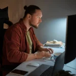 Homem trabalhando no escritório com foco no computador, ambiente de trabalho organizado e iluminação adequada, ideal para produtividade.