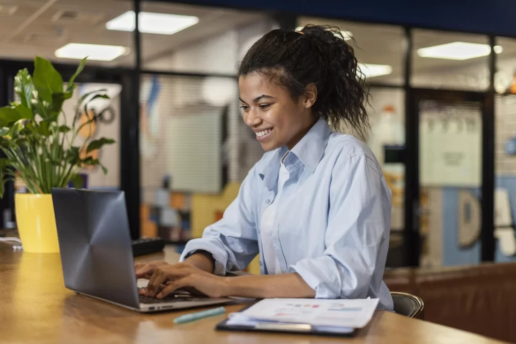 Mulher jovem e sorridente usando laptop em ambiente de escrit&oacute;rio com plantas e ilumina&ccedil;&atilde;o adequada.