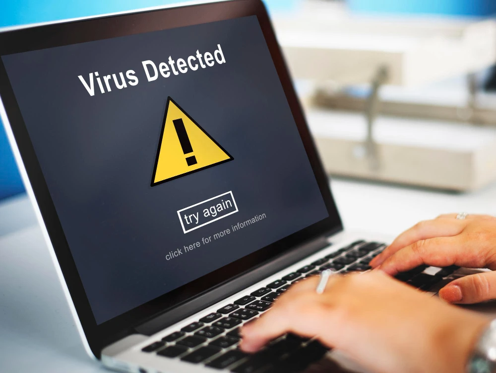 Tela de notebook exibindo alerta de “Virus Detected” com ícone de triângulo amarelo e botão “try again” enquanto uma pessoa digita no teclado, sugerindo possível infecção por malware.