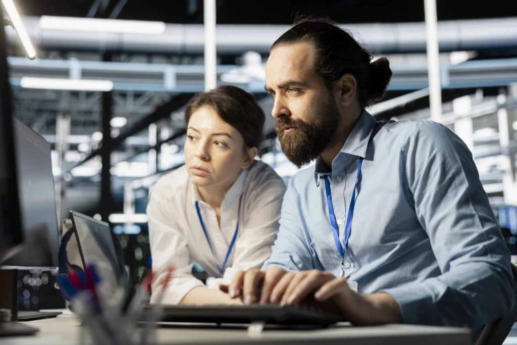 Engenheiros em uma indústria analisam informações em um monitor, enquanto um homem de barba aponta para a tela e uma mulher observa com atenção em ambiente de fábrica automatizada.