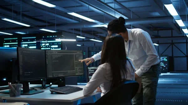 Equipe em um centro de dados observa telas de servidores e estações de trabalho, enquanto um profissional aponta para o monitor em um ambiente industrial com iluminação azul e luzes de teto