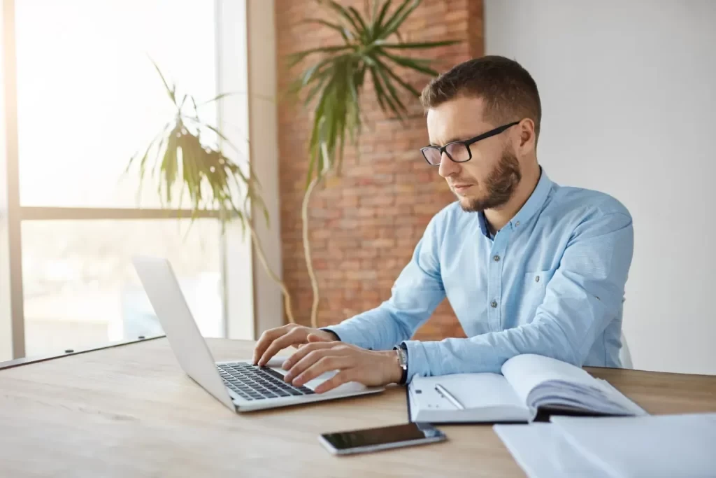 Homem com óculos digitando no notebook em um escritório moderno, usando camisa azul, com documentos abertos e celular sobre a mesa, ao lado de janela com luz natural e plantas
