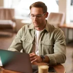 Homem trabalhando em um escritório, usando óculos e notebook sobre a mesa, com celular na mão e ambiente corporativo ao fundo desfocado, em foto colorida.