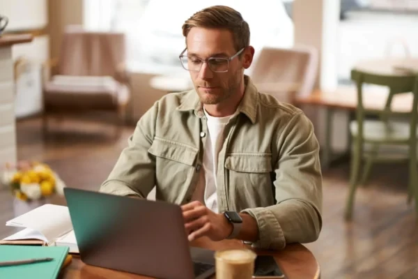 Homem trabalhando em um escritório, usando óculos e notebook sobre a mesa, com celular na mão e ambiente corporativo ao fundo desfocado, em foto colorida.
