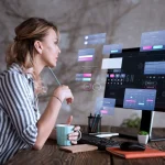 Mulher em ambiente de trabalho, ao lado de um computador com telas de interface e gráficos, enquanto escreve com caneta e toma café, sugerindo análise de dados e tecnologia.