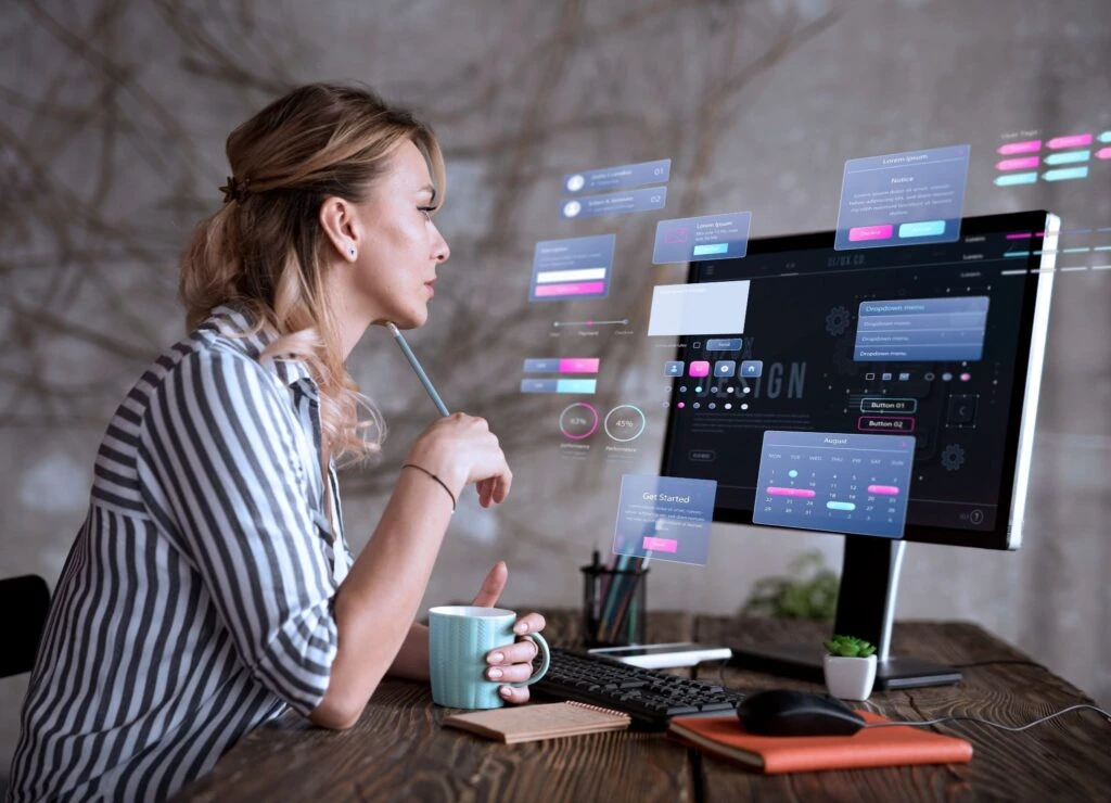 Mulher em ambiente de trabalho, ao lado de um computador com telas de interface e gráficos, enquanto escreve com caneta e toma café, sugerindo análise de dados e tecnologia.