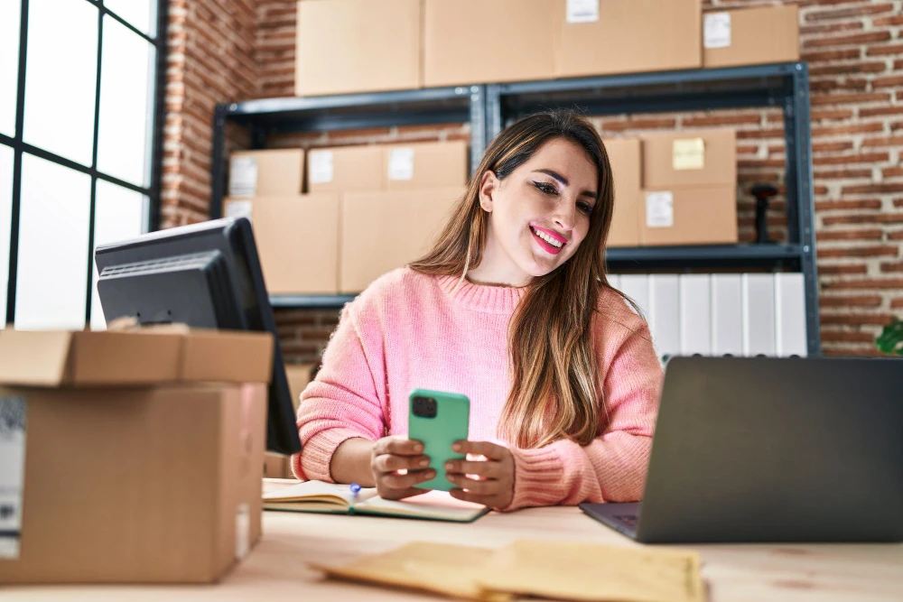 Mulher sorridente em um escrit&oacute;rio/galp&atilde;o de log&iacute;stica, embalando encomendas e usando o celular e o laptop sobre uma mesa com caixas de papel&atilde;o ao fundo. Atividade de e-commerce.