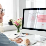 Mulher trabalhando em um escritório moderno, ao lado de um monitor de computador exibindo um site com a seção “START UP”, com mesa organizada e flores ao fundo.