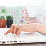 Pessoa digitando em um notebook branco sobre uma mesa, com jardim de suculentas ao fundo e fundo colorido. Mãos e teclado em foco, em ambiente de escritório ou estudo.