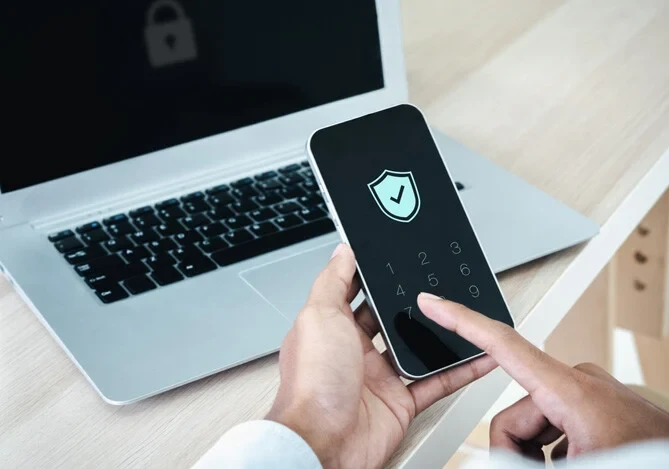 Pessoa usando smartphone com &iacute;cone de verifica&ccedil;&atilde;o e menu de seguran&ccedil;a, enquanto o notebook est&aacute; aberto em uma mesa de escrit&oacute;rio, sugerindo autentica&ccedil;&atilde;o e prote&ccedil;&atilde;o de conta.