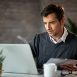 Homem trabalhando em escritório, usando notebook e analisando documentos em uma mesa com xícara e planta decorativa ao fundo, em ambiente corporativo com paredes de tijolo.