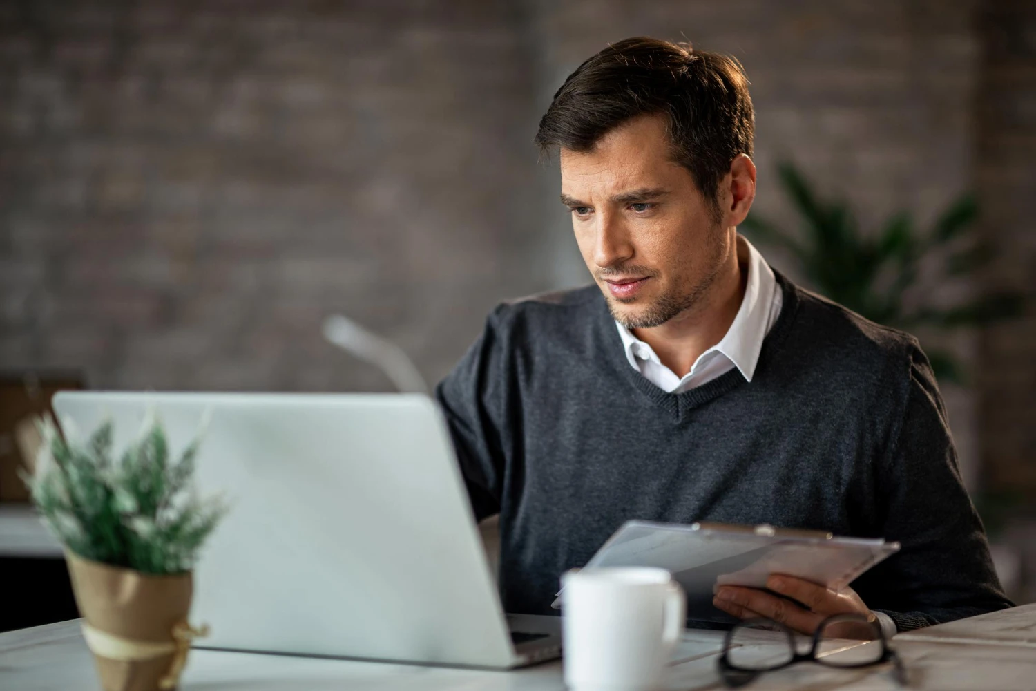 Homem trabalhando em escritório, usando notebook e analisando documentos em uma mesa com xícara e planta decorativa ao fundo, em ambiente corporativo com paredes de tijolo.